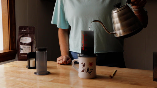 Person pouring hot water into an AeroPress coffee maker on a wooden kitchen island.