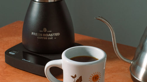 A diner mug in front of an electric kettle beside a scale and coffee carafe.