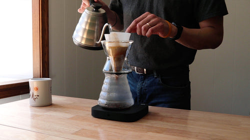 Person pouring hot water over coffee grounds in a Chemex Funnex pour over dripper on a scale.