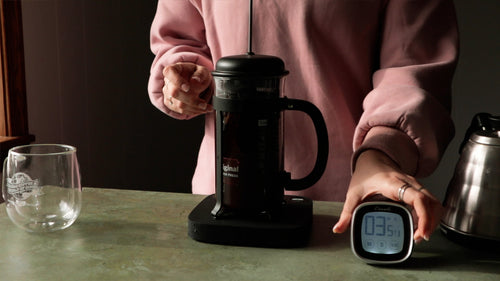 A French press being brewed on a scale by someone in a pink sweatshirt setting a timer.