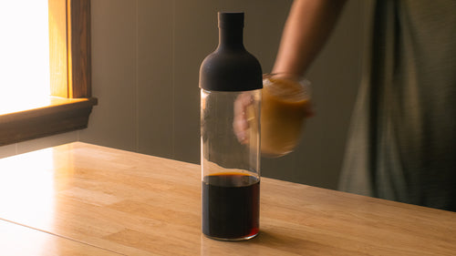A Hario Cold Brew Bottle with coffee in the bottom on a kitchen island with a hand pulling away a glass of cold brew in the background.