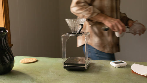A person walking behind a Hario V60 Drip Station on a table next to a coaster, tier, and hot water carafe.