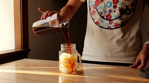 A person pouring cold brew coffee over ice in a Mason jar.