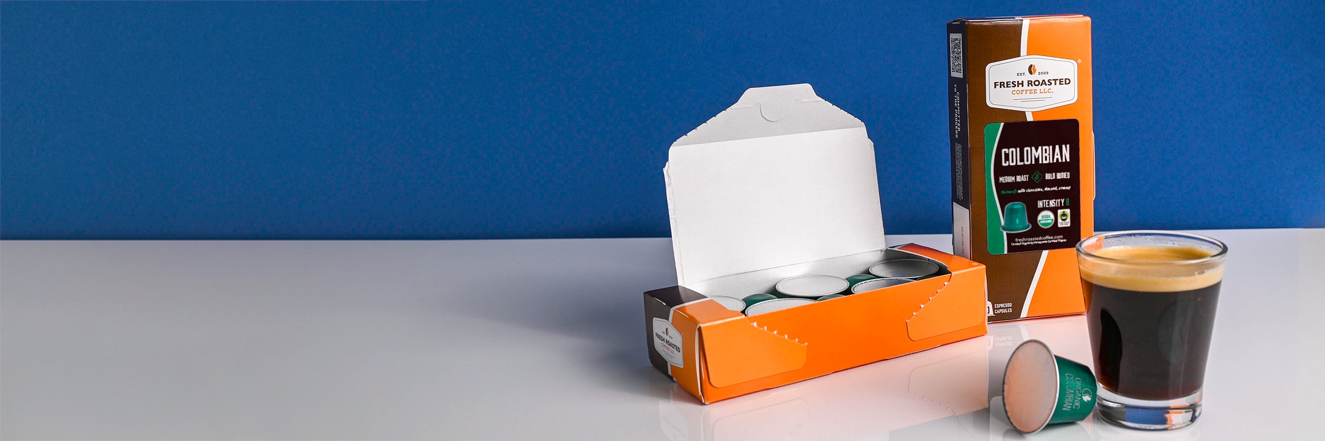 Organic Colombian espresso capsule carton next to a shot of espresso and capsule.