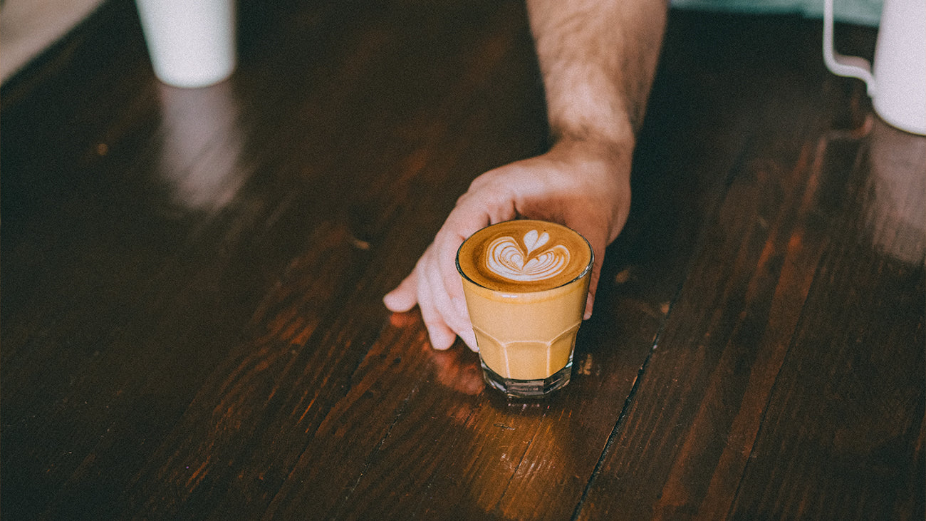 A barista sliding a flat white across a wood-top coffee bar.