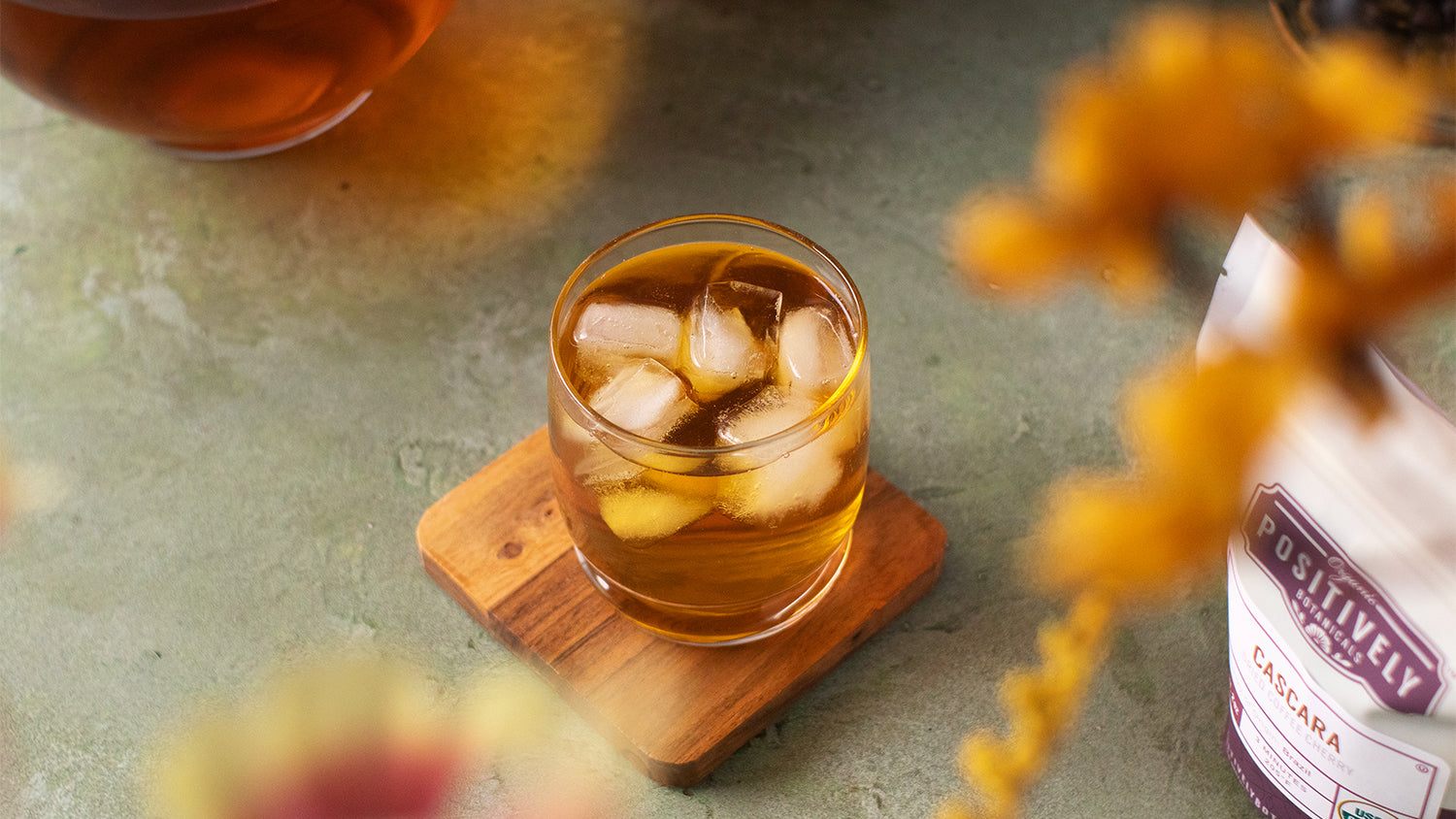 A glass of iced cascara on a coaster on a green background with florals in the foreground.