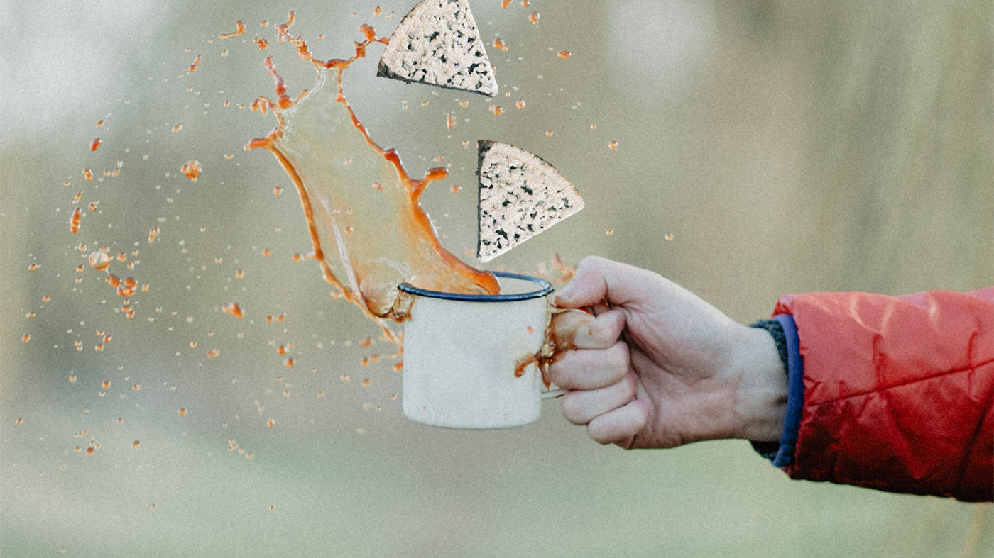 Two wedges of cheese falling into an enamel mug splashing coffee, held up by a hand.
