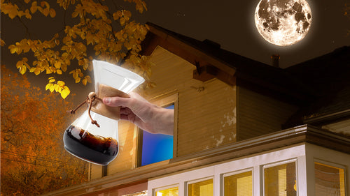 A hand extending a Chemex coffee maker out of a second-story window at night with the sun high in the sky.