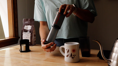 Person holding an AeroPress upside down on a kitchen counter with filters, a coffee bag, diner mug, scale, and kettle nearby.