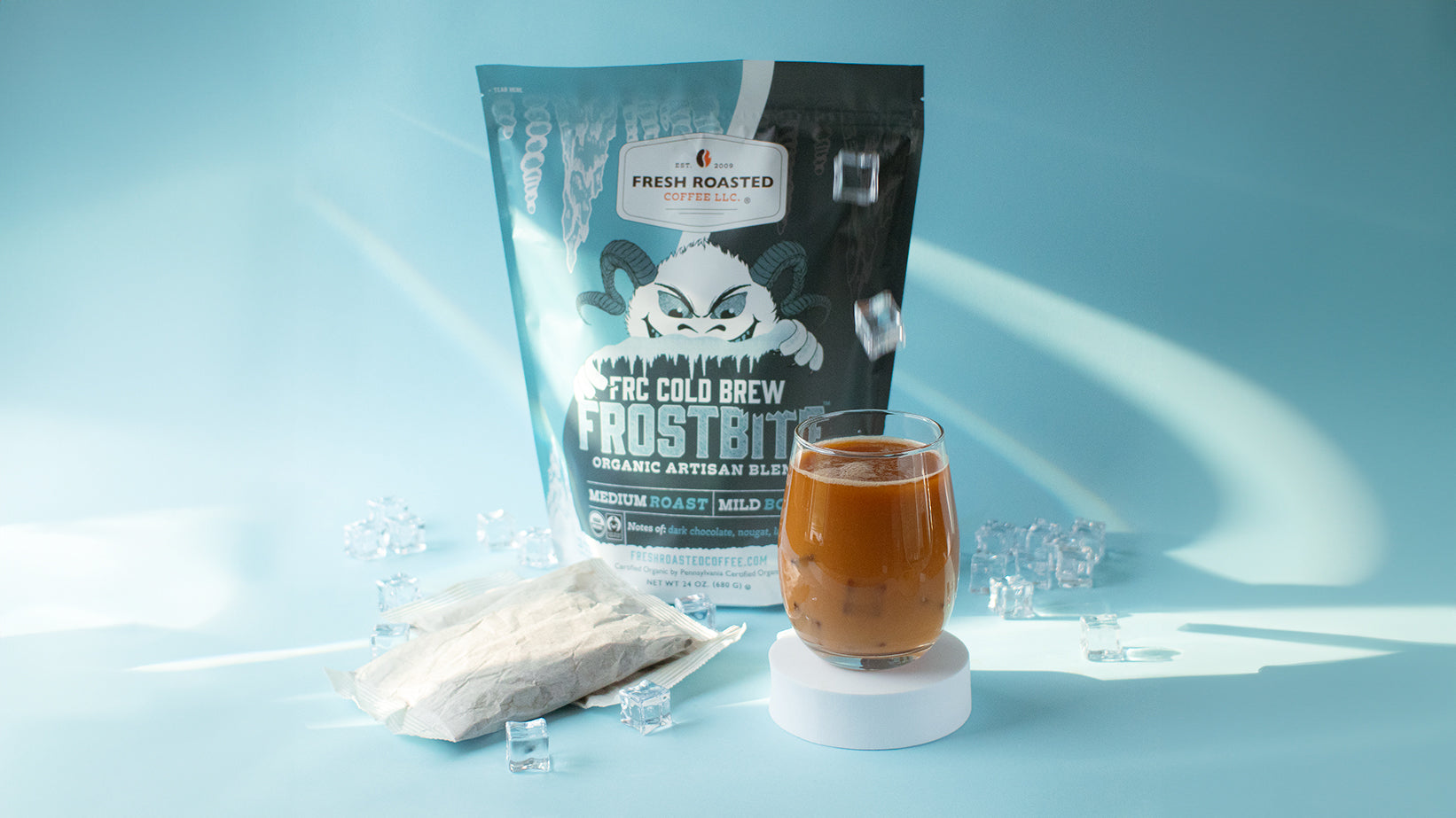 A fully printed, blue-monochrome bag on a white platform behind two cold brew filter packs and a glass of cold brew on another platform, all on a blue background.