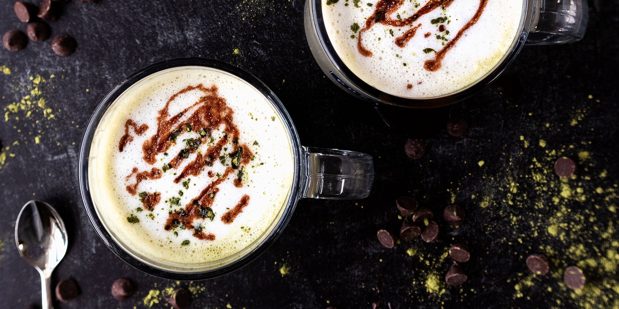 Two minty, chocolatey matcha lattes pictured from above, with matcha powder, chocolate chips, and smalls spoons scattered about.