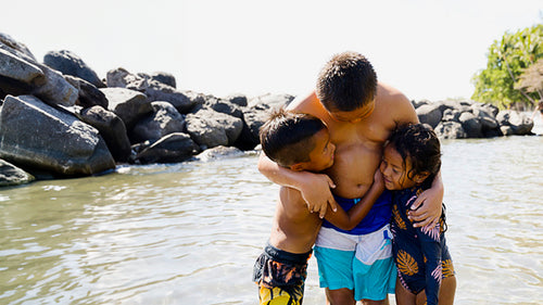 A group of Hawaiian kids hugging while swimming in the ocean near large boulders.