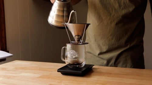 Coffee person pouring water from a gooseneck kettle into a stainless-steel dripper seated atop a double-walled mug on top of a black scale.