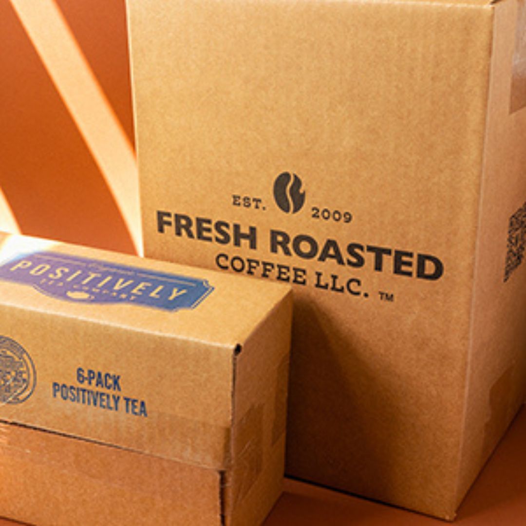 A small cardboard shipping box with a blue Positively Tea logo, sitting on it's side in front of a larger cardboard shipping box standing upright, with a black Fresh Roasted Coffee LLC logo on it.