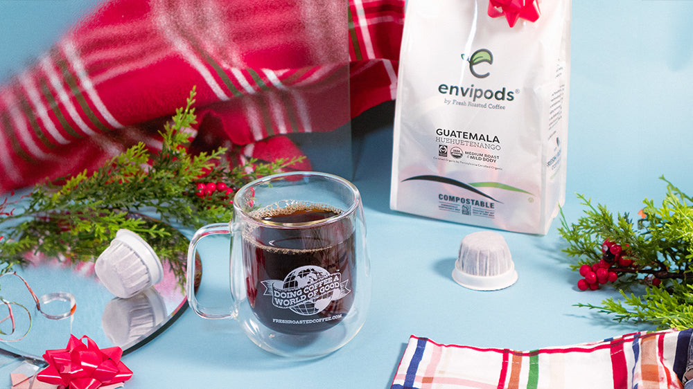 A double-walled coffee mug, a bag of compostable pods, and wintry foliage, bows, and napkins on a blue background.