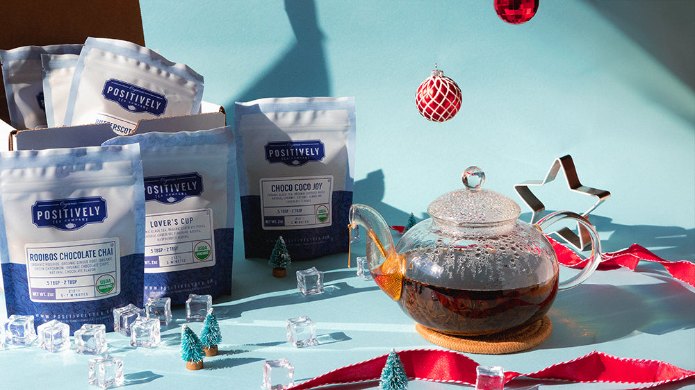 A full tea pot on a blue background, surrounded by ornaments, ribbons, ice, and loose-leaf bagged tea.