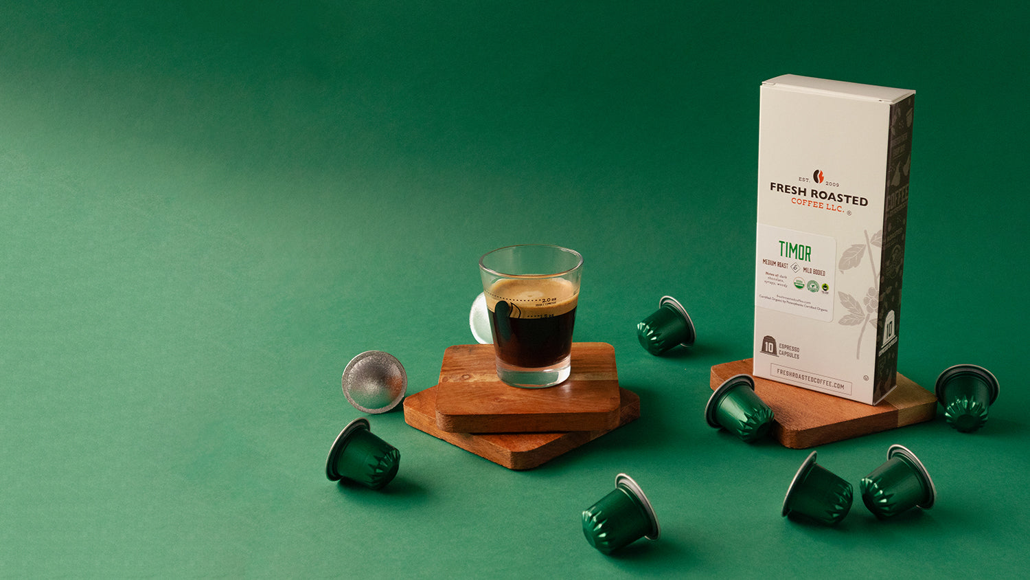 Tall white box next to a shot of espresso surrounded by green espresso capsules, all on a green background.