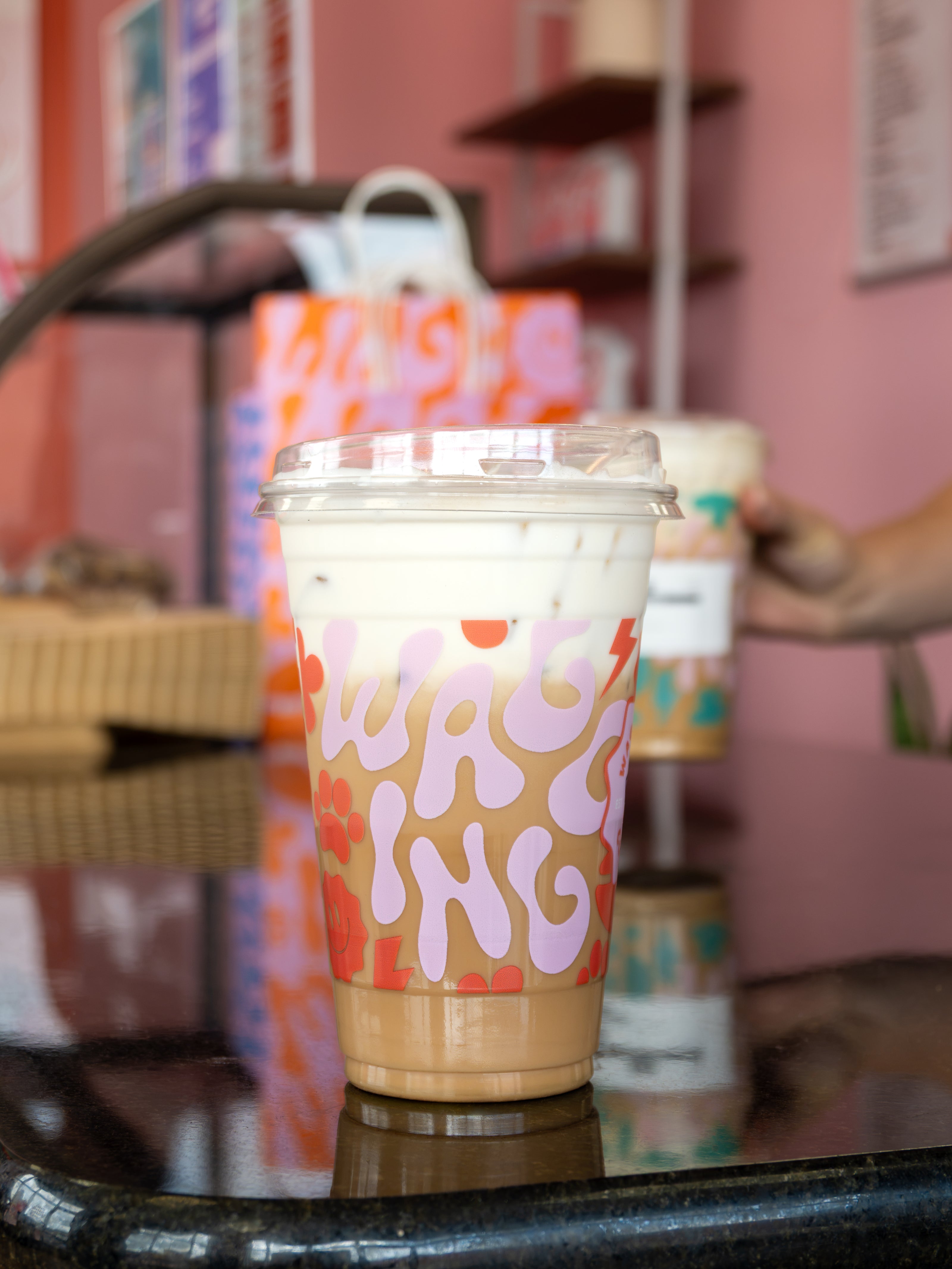 A clear plastic takeaway coffee cup with a clear lid sits on a dark table in a cafe. The cup has a pink and orange abstract pattern with the word 'WAGGING' in large pink letters.