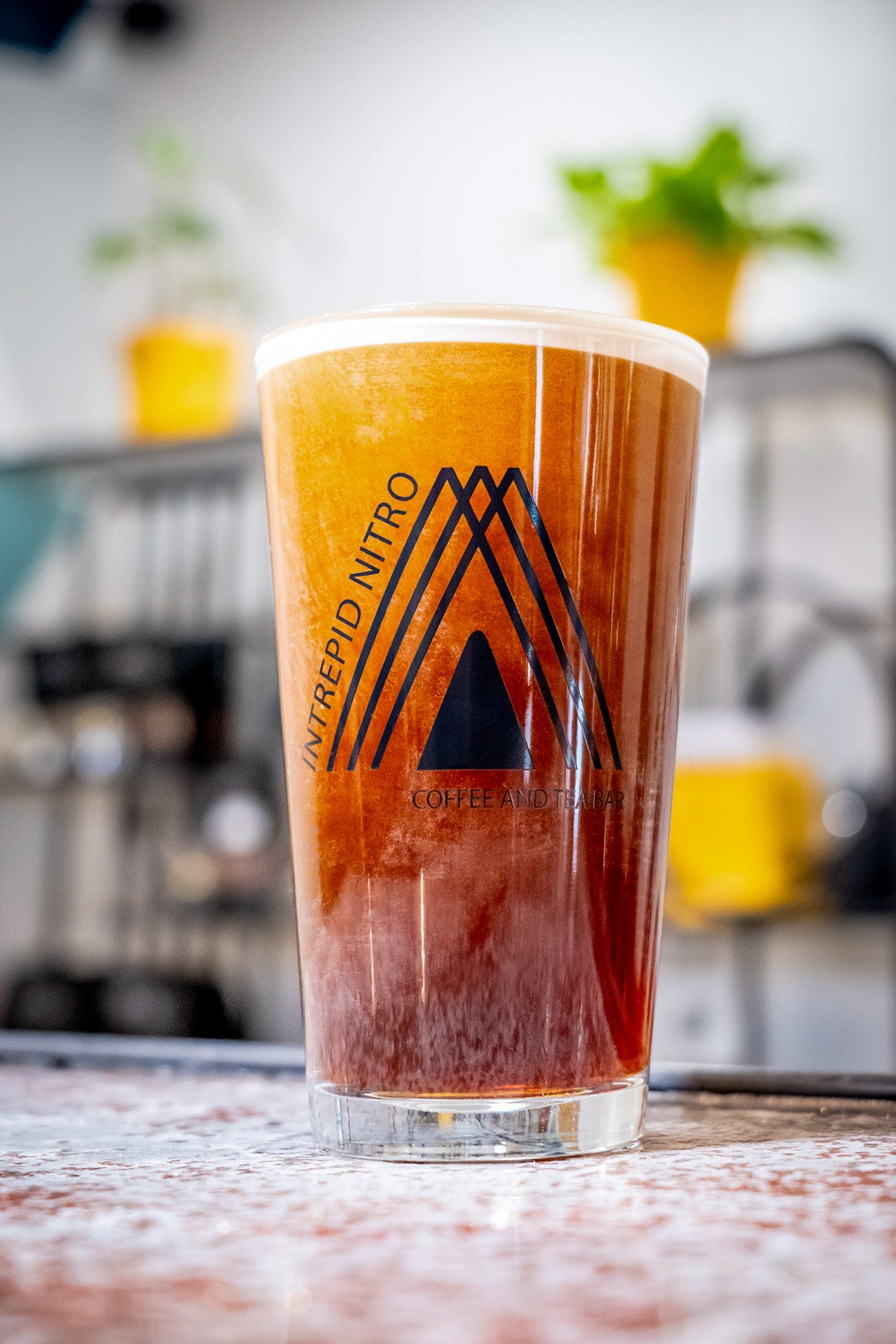 A clear pint glass with a black printed logo, filled with an amber-brown iced coffee and topped with white foam, set on a countertop in a blurred cafe-like environment.