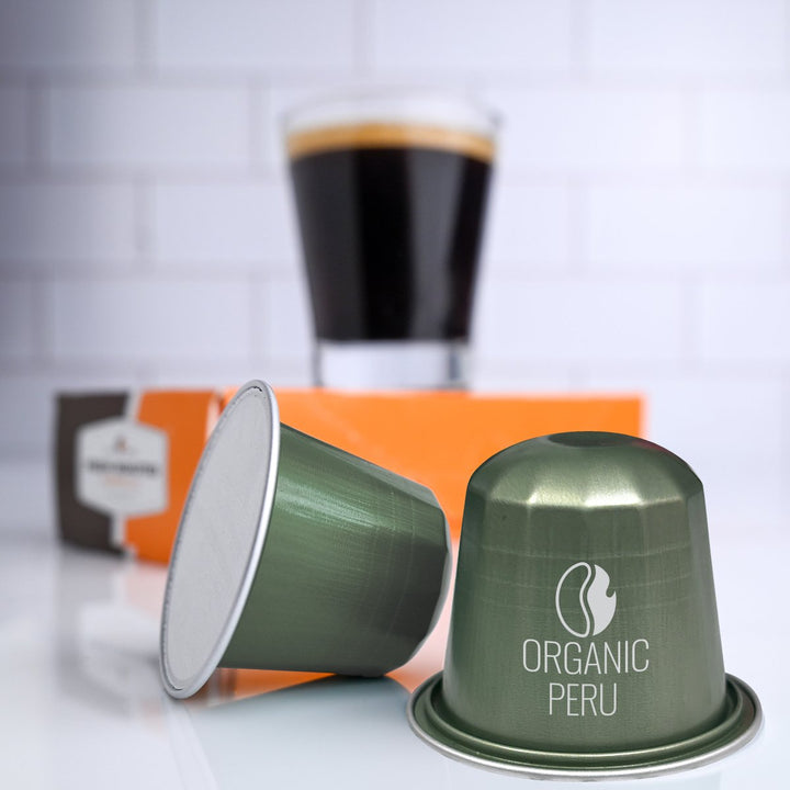 Fresh Roasted Coffee Peru Organic Half-Caf Espresso Capsules placed beside a glass of freshly brewed espresso, highlighting smooth orange and chocolate flavors.