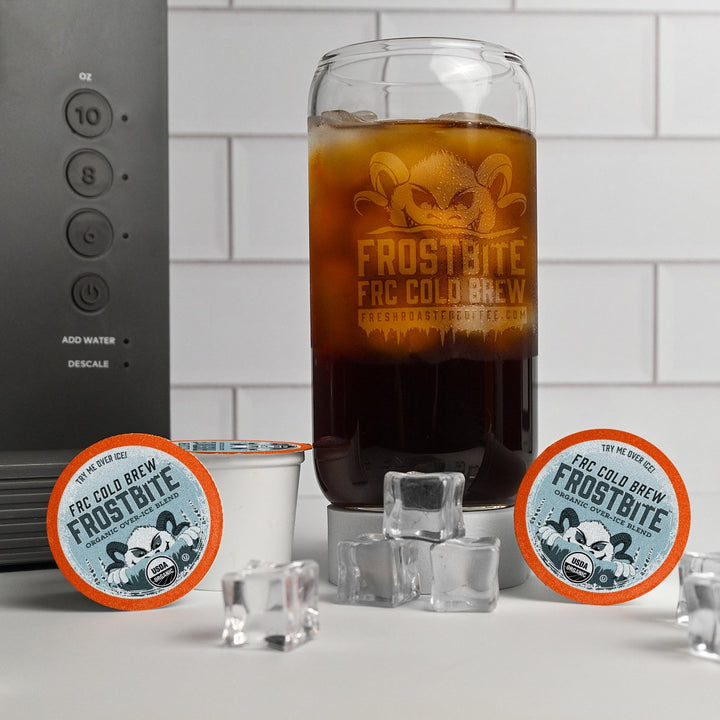 Fresh Roasted Coffee Frostbite Cold Brew served over ice in a glass, surrounded by single-serve pods, highlighting smooth chocolate and nougat flavors.
